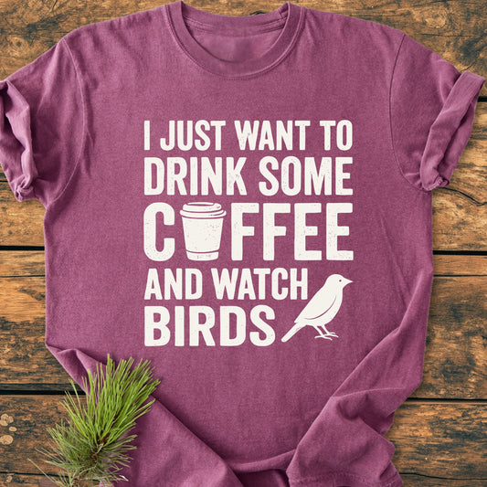 Heathered plum T-shirt 1405 Drink Coffee And Watch Birds Base with white lettering, coffee cup icon, and songbird silhouette, laid on wood. Pine sprig accents the relaxed fit, cotton fabric.