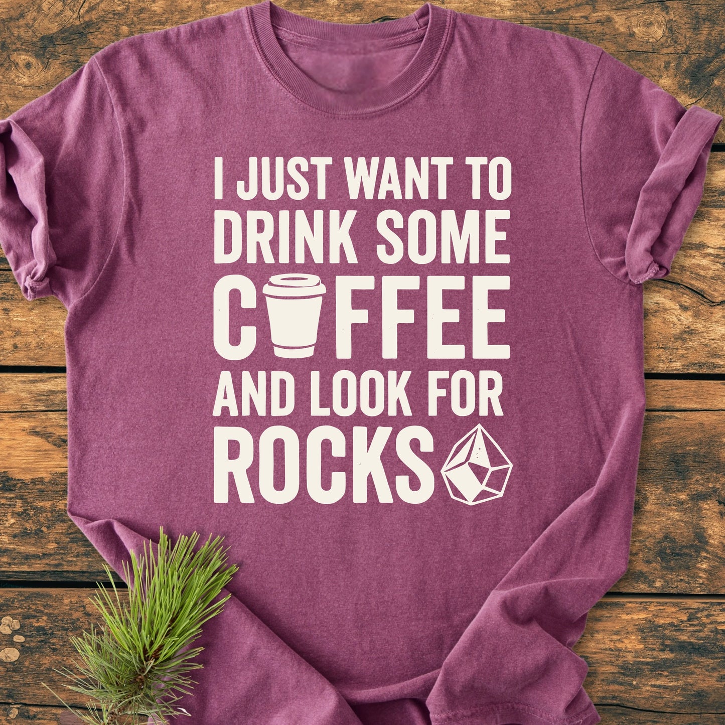 Heather-purple Comfort Colors 1717 T-shirt reading I JUST WANT TO DRINK SOME ☕️ COFFEE AND LOOK FOR ROCKS 💎, featuring a coffee cup and gemstone graphic, laid flat on wood with a pine branch.