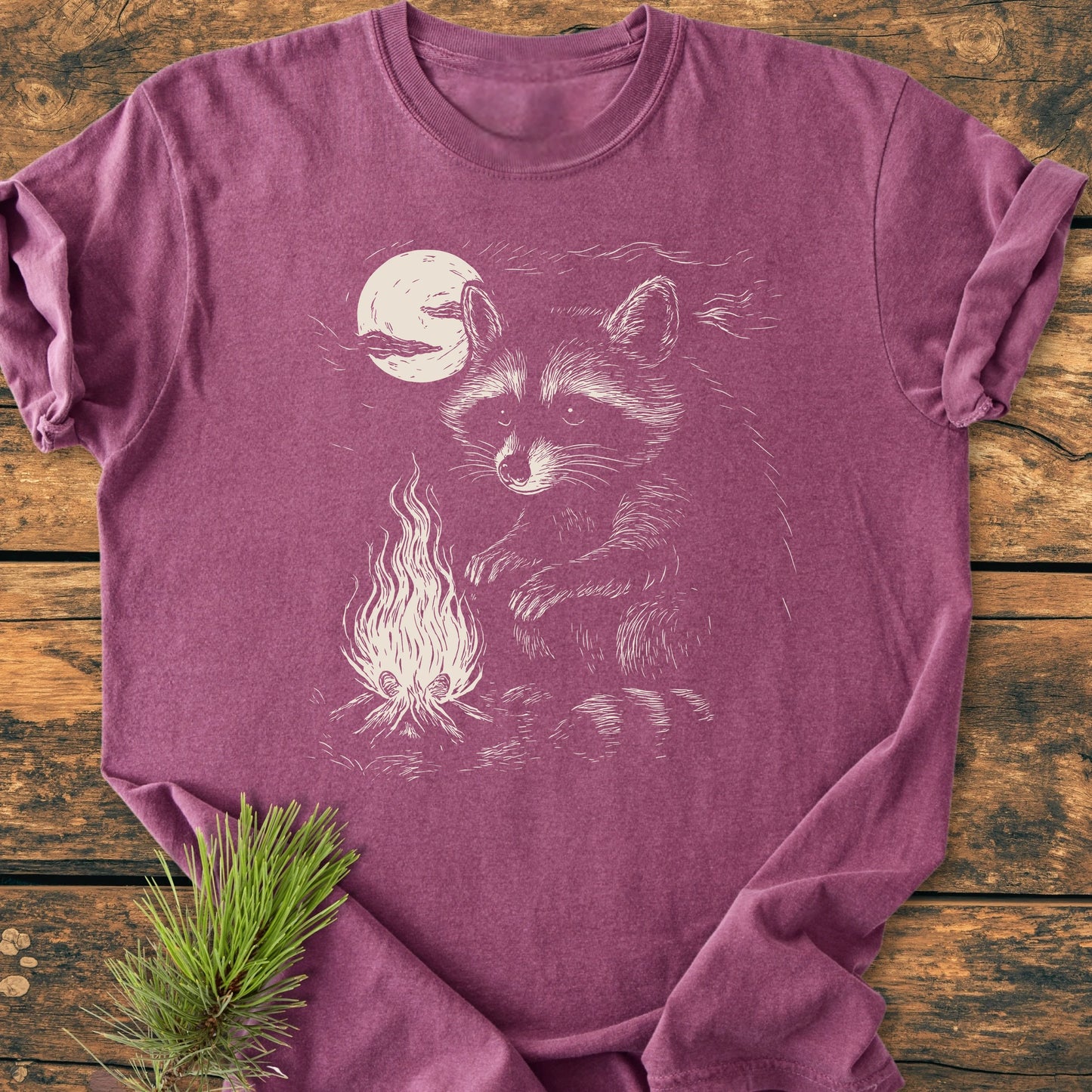 1409 Moonlit Campfire Raccoon Base T-shirt features a raccoon by campfire under full moon in off-white line art, laid on rustic wood with pine sprig, enhancing the outdoor aesthetic.