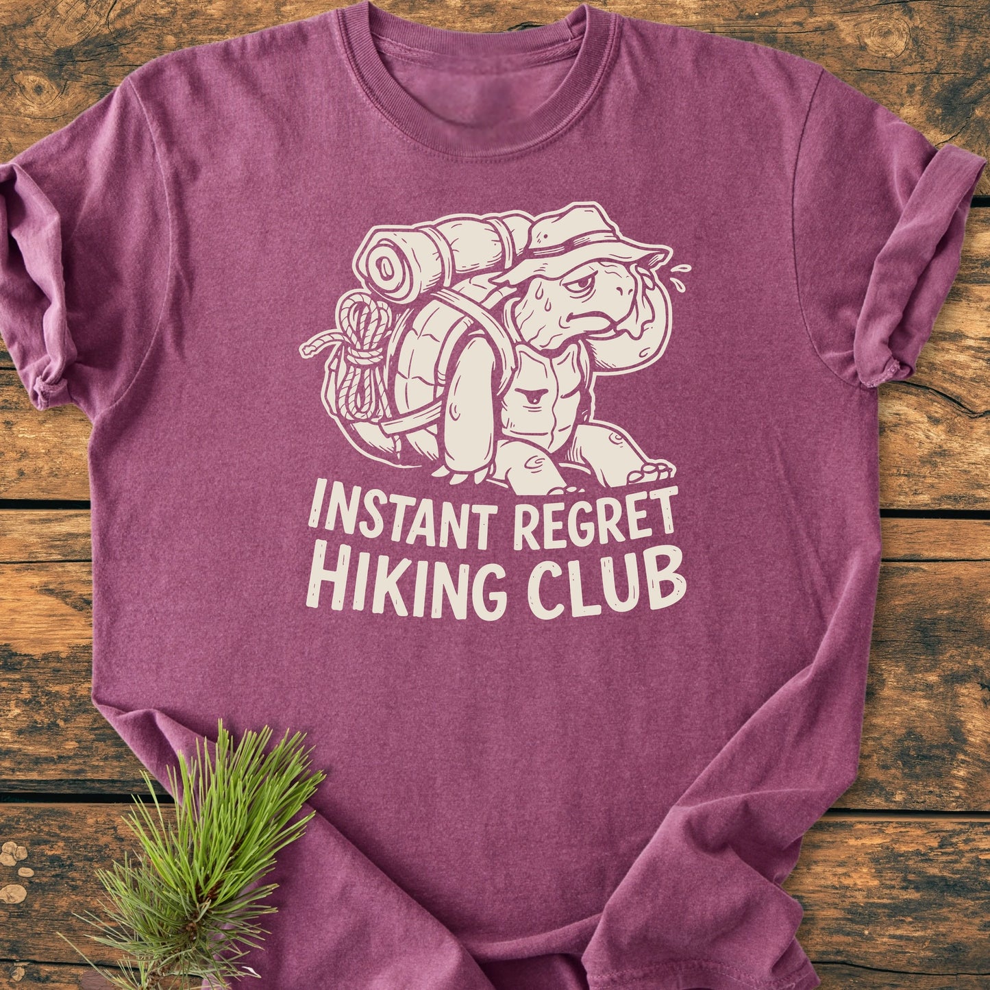 Heathered purple 1430 Instant Regret Hiking Club T-shirt featuring a cartoon hiking tortoise, relaxed unisex fit, vintage wash, and durable construction, laid flat on a wood surface.