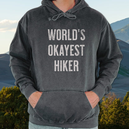 Person wearing World's Okayest Hiker Comfort Colors Hoodie with hands in kangaroo pocket, showcasing relaxed fit and vintage look against sand dunes backdrop.