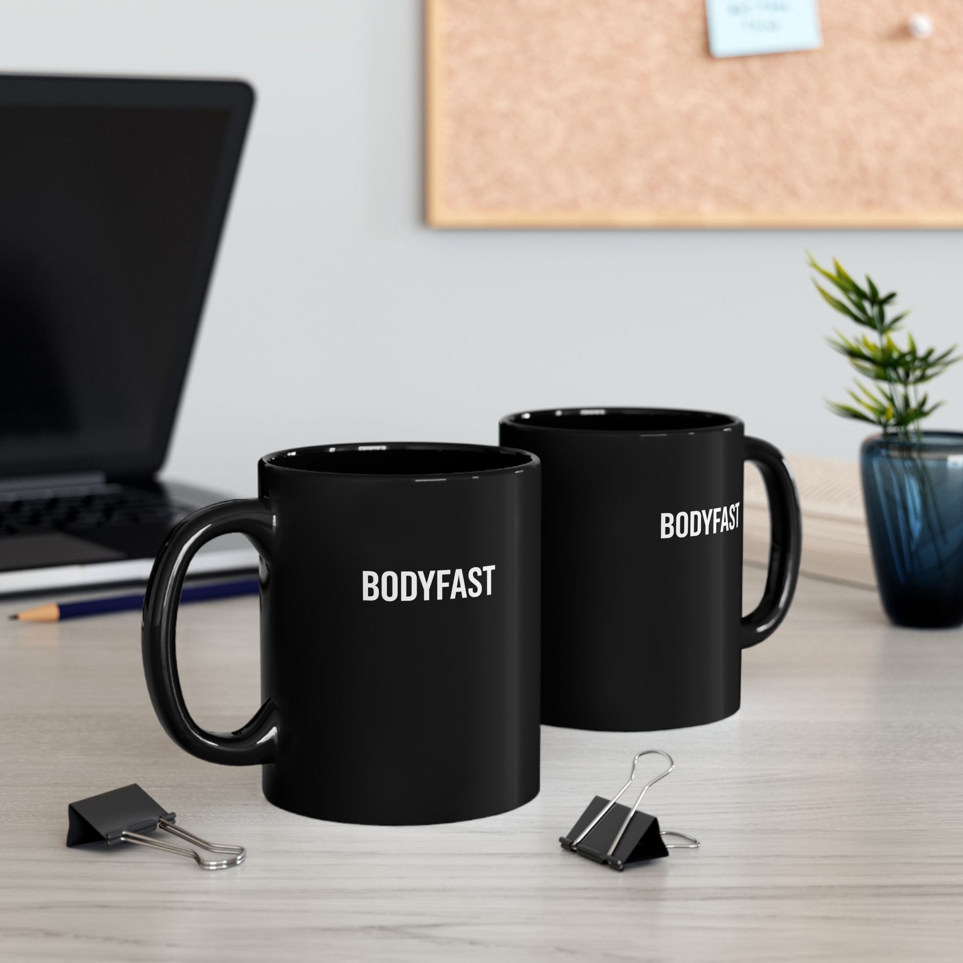 BODYFAST Ceramic Mug: Two glossy black mugs with BODYFAST in white on a light-wood desk, surrounded by binder clips and a pencil. A laptop, cork board, and plant complete the setting.