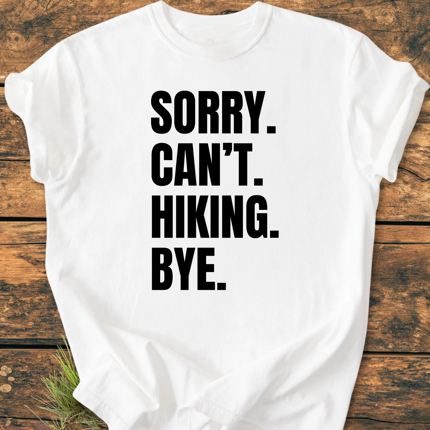 Plain white T-shirt on a wooden surface, featuring SORRY. CAN’T. HIKING. BYE. in bold black text. Sprigs of greenery add an outdoorsy touch, complementing the 1413 Sorry. Can't. Hiking theme.