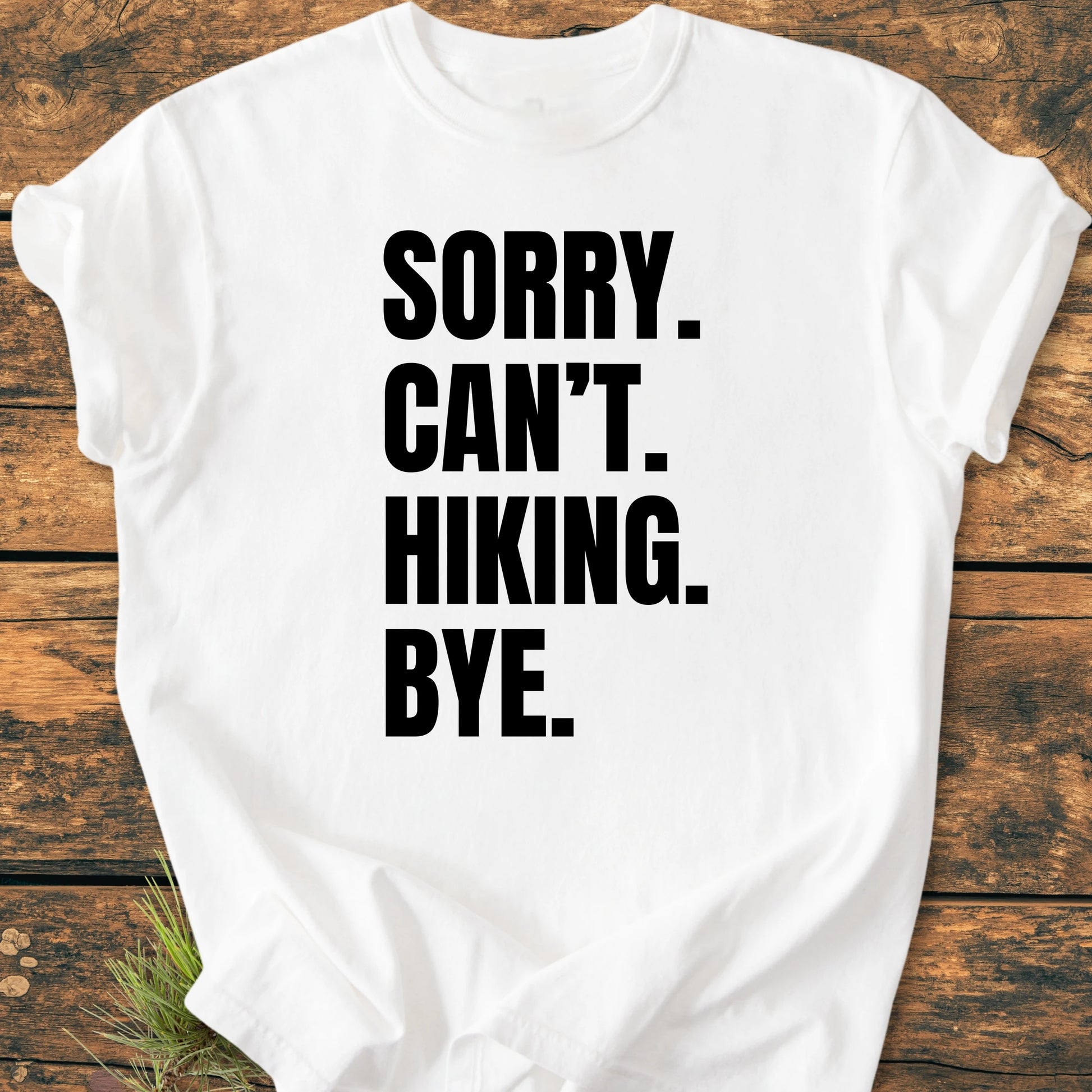 Plain white T-shirt on a wooden surface, featuring SORRY. CAN’T. HIKING. BYE. in bold black text. Sprigs of greenery add an outdoorsy touch, complementing the 1413 Sorry. Can't. Hiking theme.