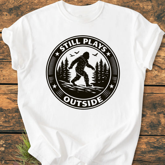 1430 Still Plays Outside Bigfoot T-shirt featuring a Sasquatch graphic amidst a pine forest. Relaxed unisex fit, vintage wash, soft garment-dyed cotton, ideal for outdoor enthusiasts seeking comfort and style.