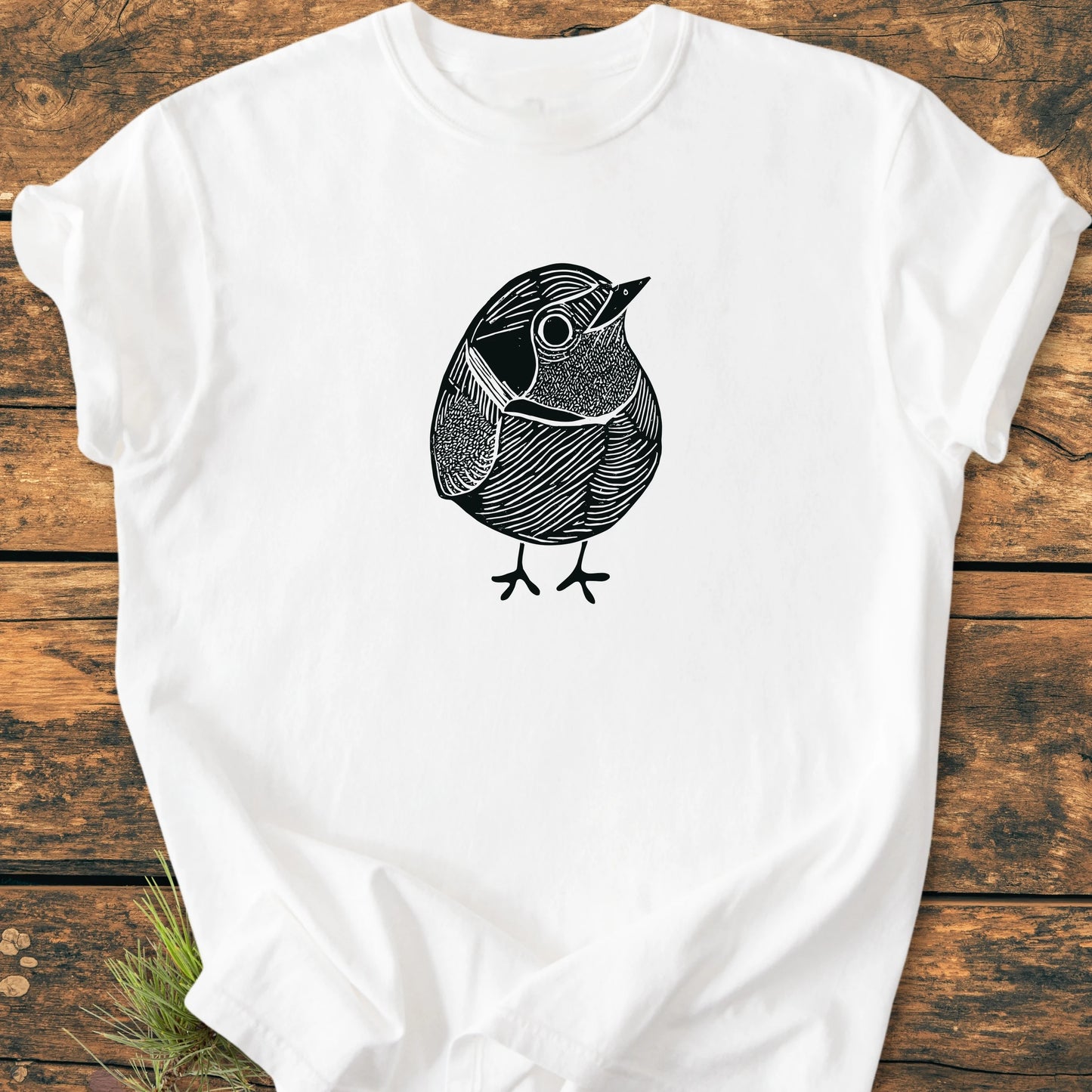 White T-shirt featuring 1433 Little Robin design, a black line-drawing of a small bird, laid flat on wooden planks with pine needles visible, highlighting its relaxed unisex fit and vintage wash.