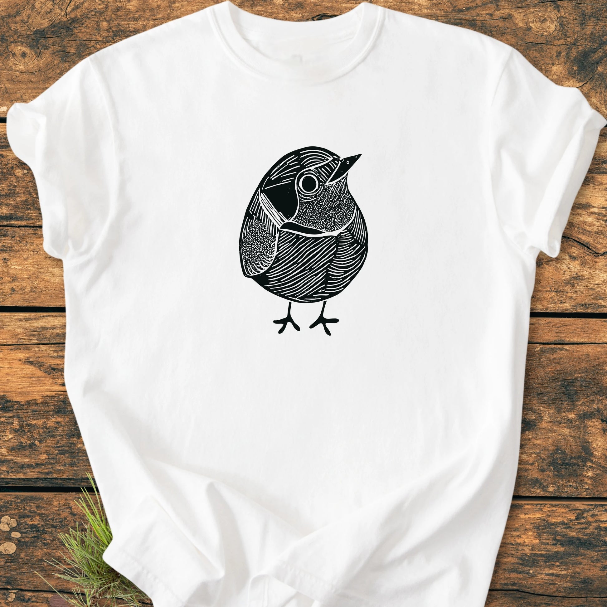 White T-shirt featuring 1433 Little Robin design, a black line-drawing of a small bird, laid flat on wooden planks with pine needles visible, highlighting its relaxed unisex fit and vintage wash.
