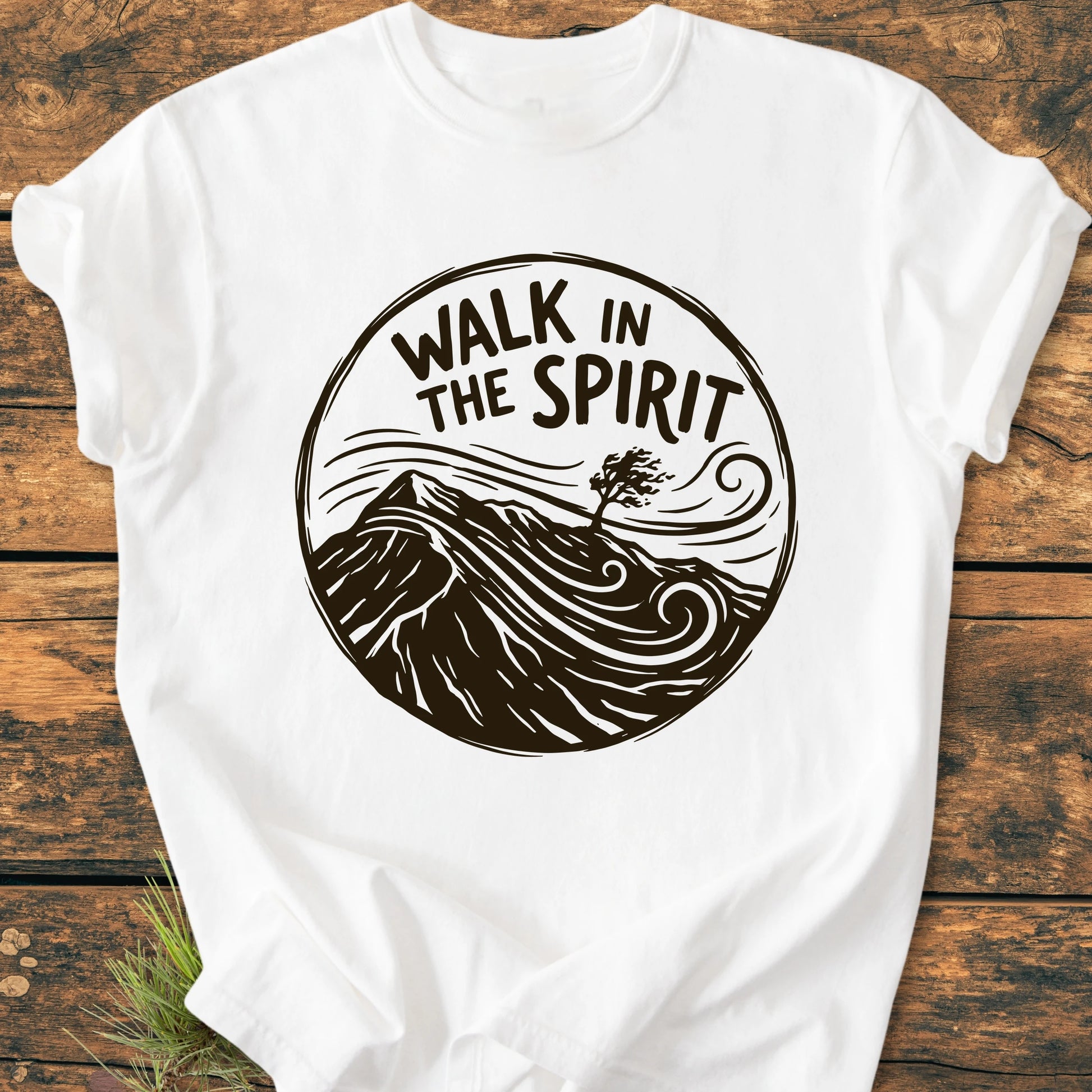 White T-shirt titled 1410 Walk In The Spirit featuring a circular graphic with mountains and a tree, set against a wooden surface. Made from soft, garment-dyed, 100% ring-spun cotton.