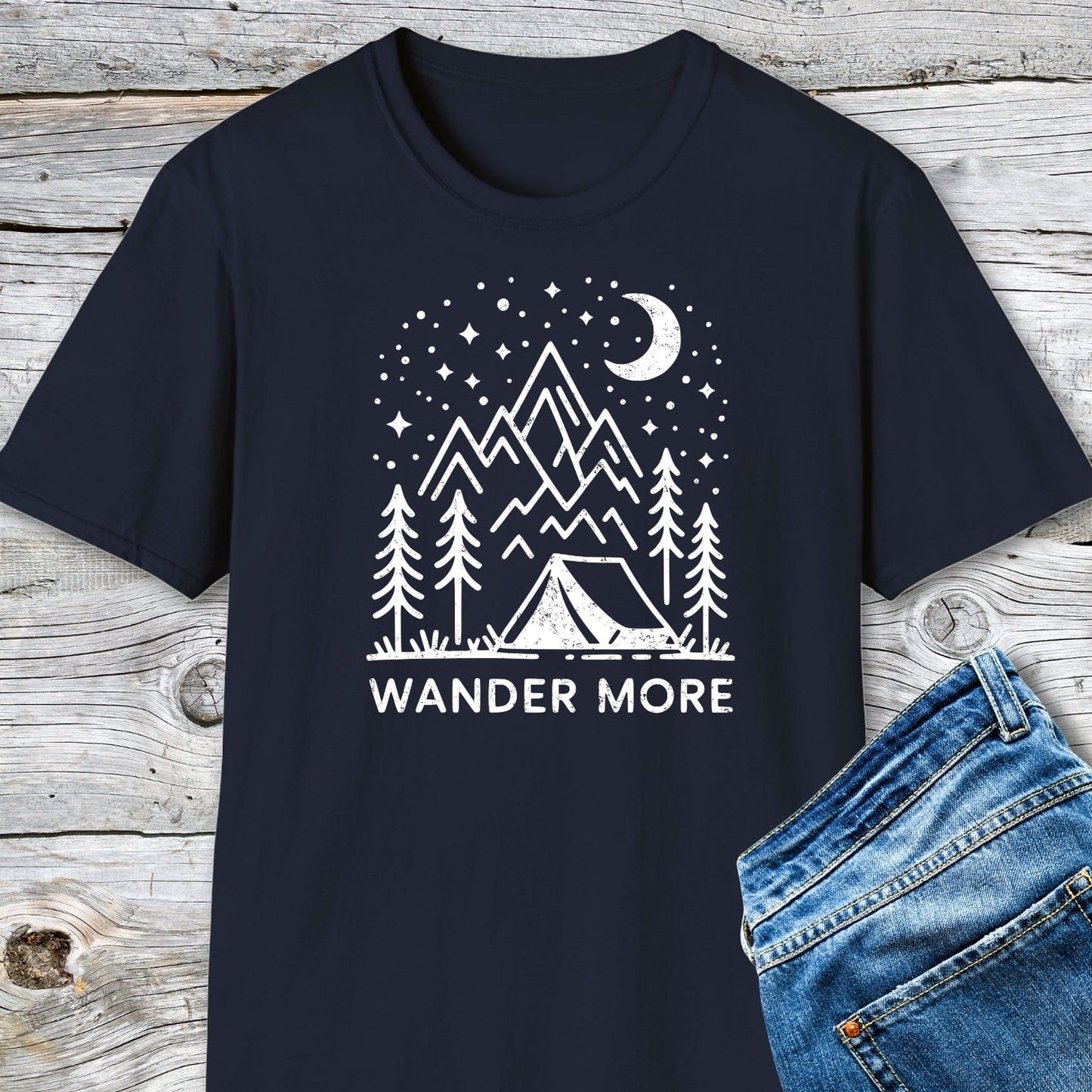 Wander More T-Shirt featuring a nighttime camping scene graphic on soft cotton, showcasing mountains, stars, and a crescent moon, laid on a wooden surface.
