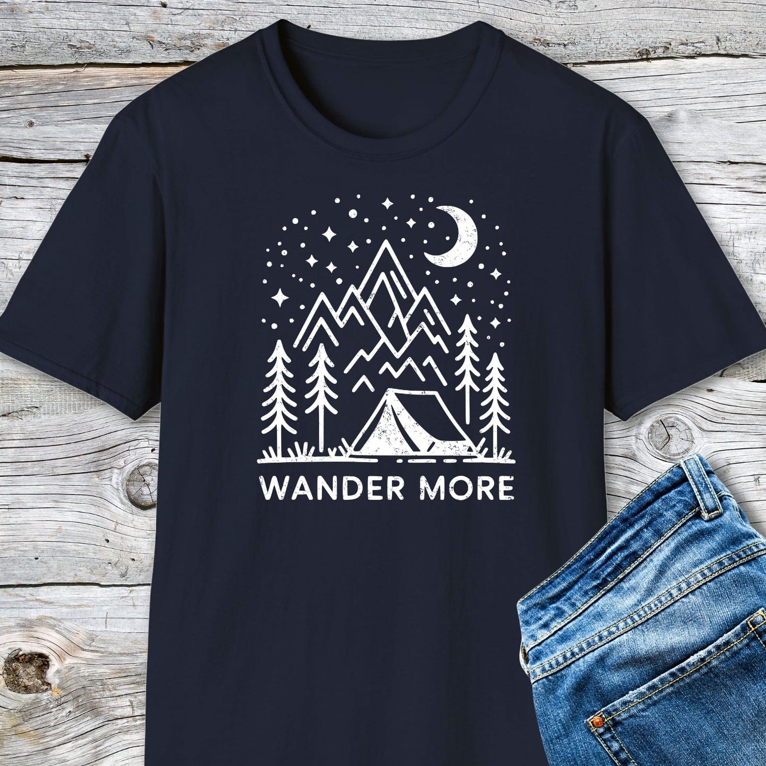 Wander More T-Shirt featuring a nighttime camping scene graphic on soft cotton, showcasing mountains, stars, and a crescent moon, laid on a wooden surface.