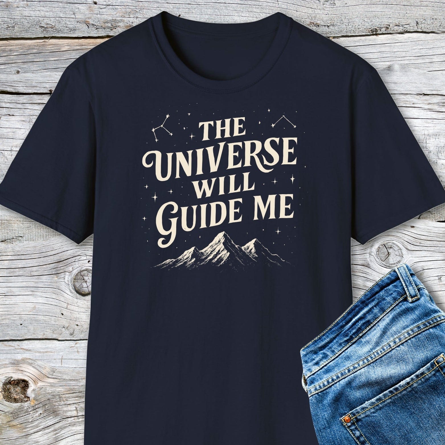 The Universe Guides Me T-Shirt: Navy-blue tee with cream text and stars, made from soft cotton for comfort, accompanied by blue jeans on a wooden surface.
