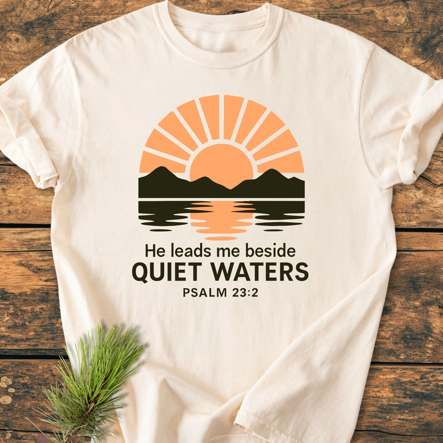 The 1424 He Leads Me T-shirt showcases a sunset mountain graphic with Psalm 23:2 text, on soft, vintage-wash fabric, designed for comfort and durability, laid on a wooden surface.