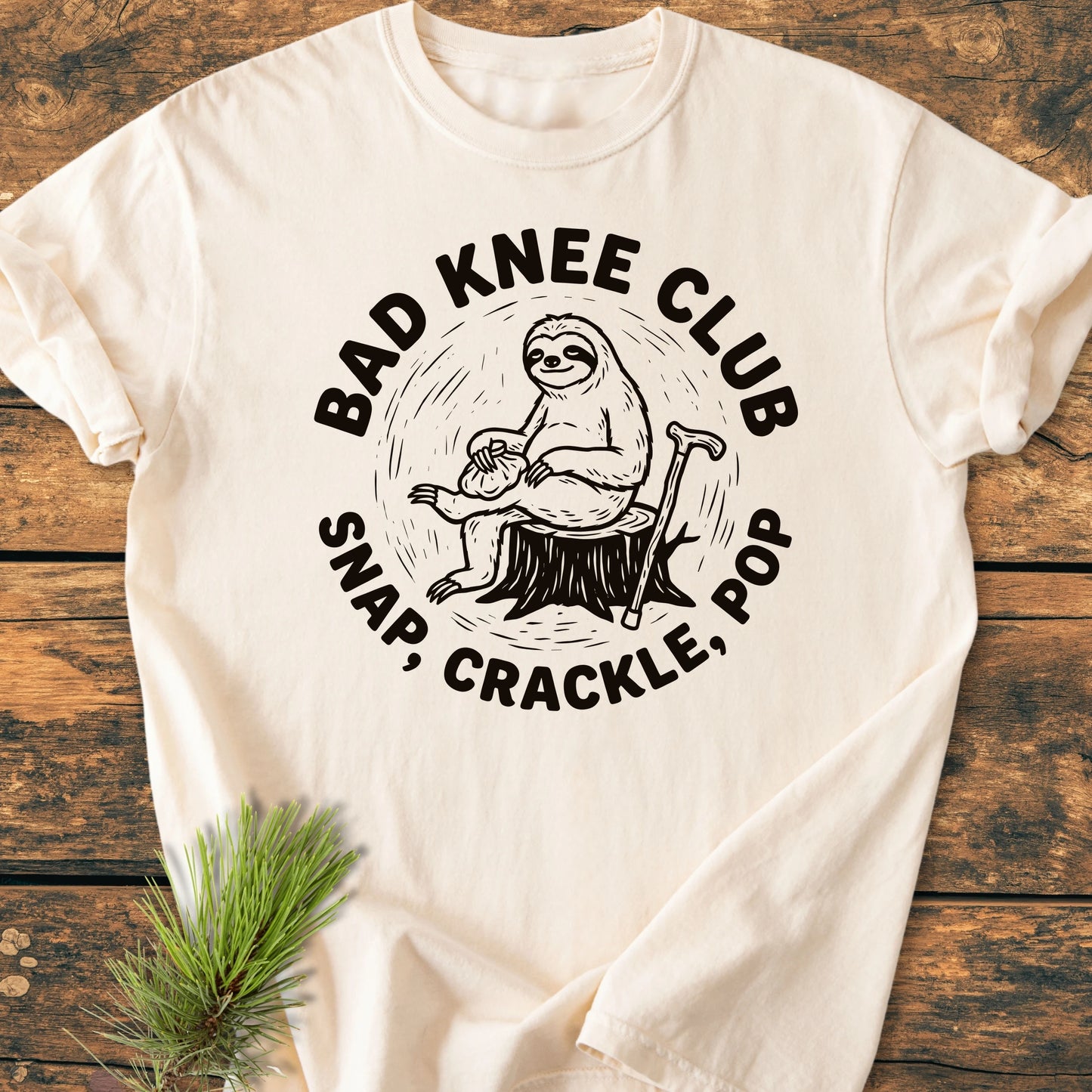 Light-colored T-shirt featuring BAD KNEE CLUB graphic of a sloth with an ice pack, showcasing a vintage wash and relaxed fit, styled on a wooden surface with a pine sprig.