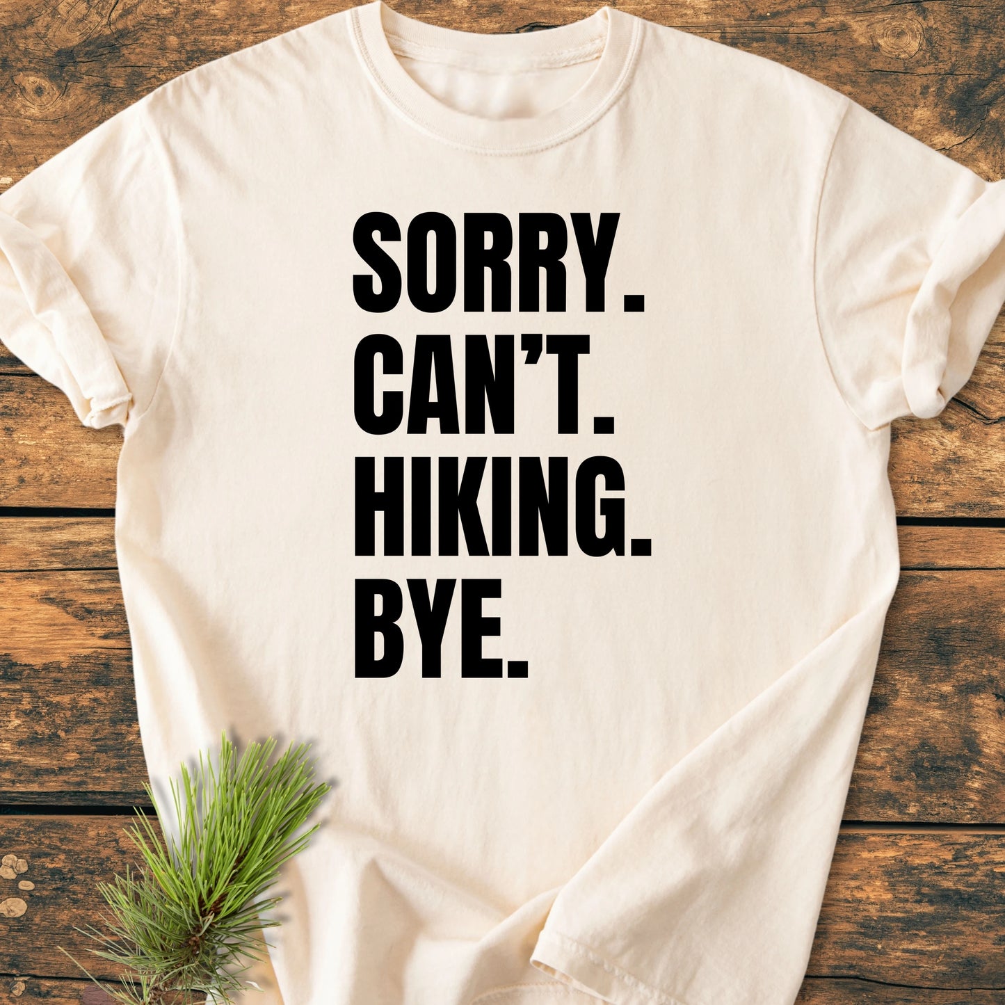 Cream-colored T-shirt with SORRY. CAN’T. HIKING. BYE. printed in black, displayed on wood. It's a vintage wash tee, soft, relaxed fit, ideal for hiking enthusiasts and everyday wear.