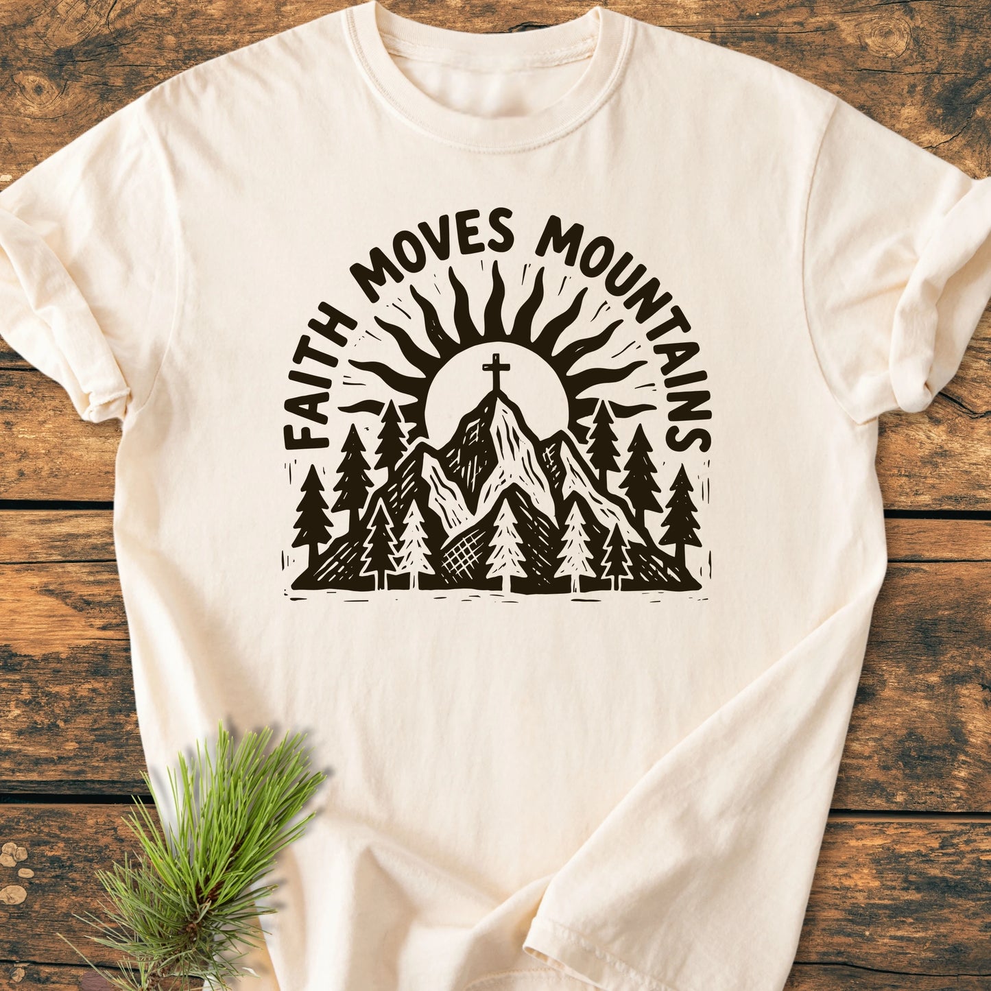 Ivory cotton T-shirt with FAITH MOVES MOUNTAINS graphic, featuring a mountain range, pine trees, sun, and cross, laid flat on wood, representing the 1409 Faith Moves Mountains design.