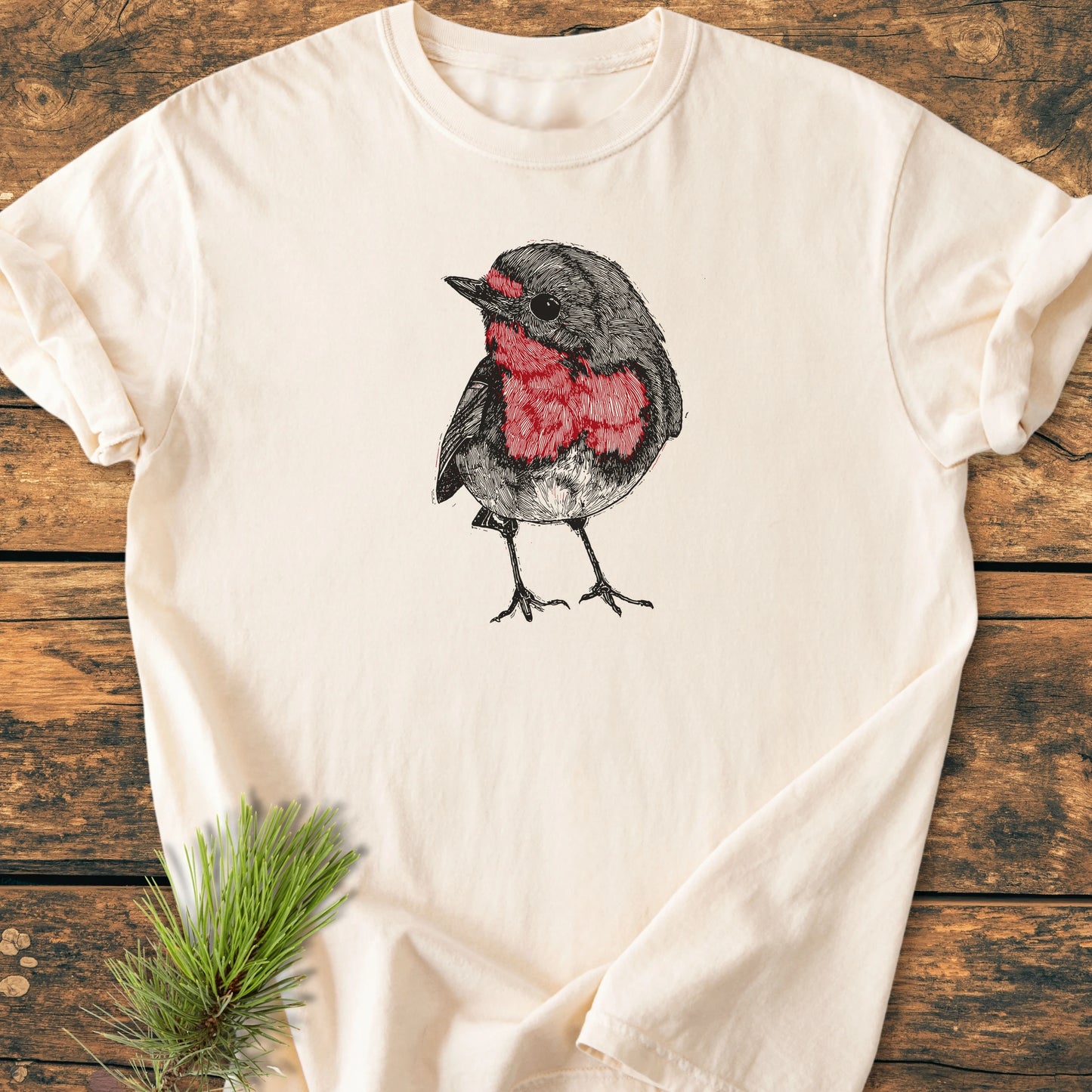 Cream-colored Cute Robin T-shirt on wood, featuring a hand-drawn robin with a red breast, highlighting its vintage wash and relaxed fit, perfect for nature enthusiasts and everyday comfort.