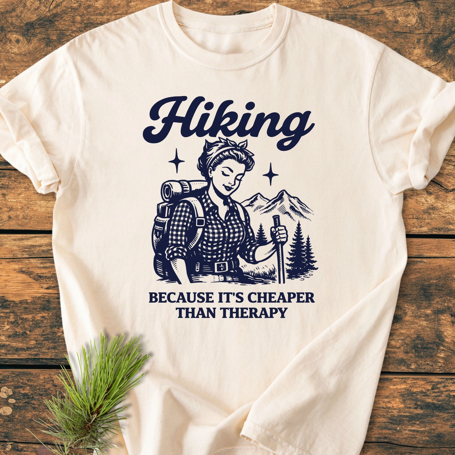 The 1407 Hiking Cheaper Than Therapy Base t-shirt features a navy graphic of a female backpacker, emphasizing durability and comfort with 100% ring-spun cotton, laid on a wooden surface.