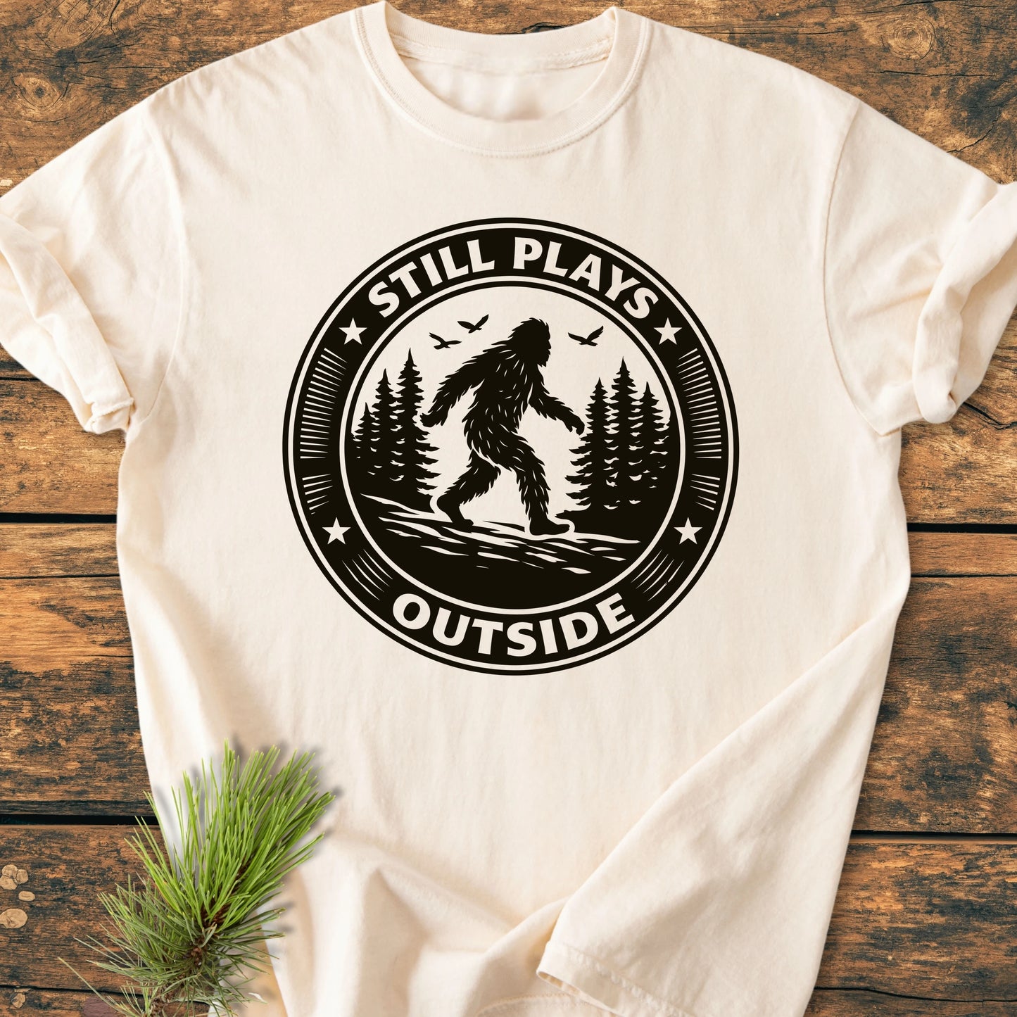 1430 Still Plays Outside Bigfoot T-shirt featuring a circular Bigfoot graphic in a forest, laid on a wooden surface with a pine sprig beside it, highlighting its vintage, relaxed fit.