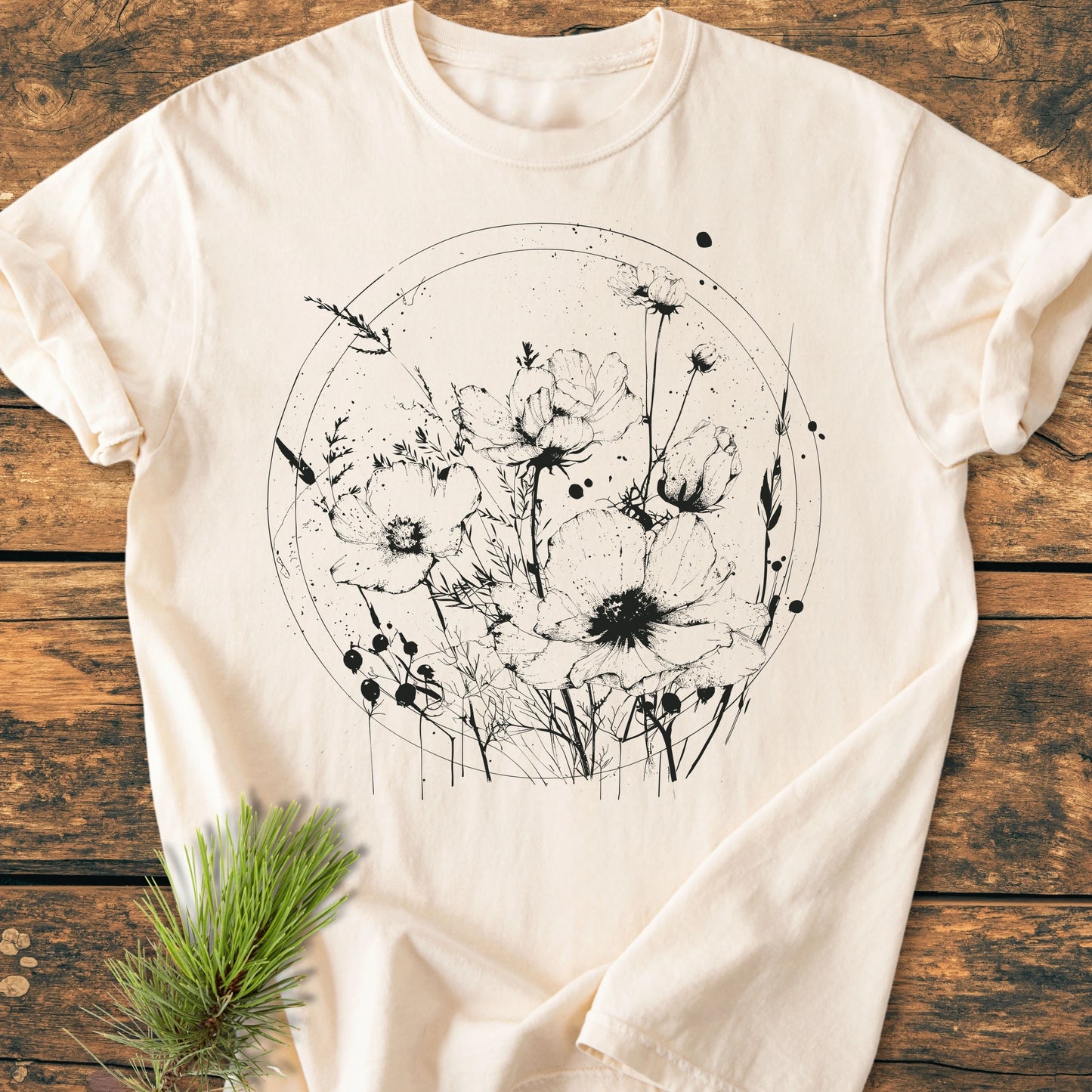 Cream-colored T-shirt 391 Wildflower Circle with black wildflower illustration on the front, styled on a wooden surface with a pine sprig, showcasing a relaxed fit and durable construction.