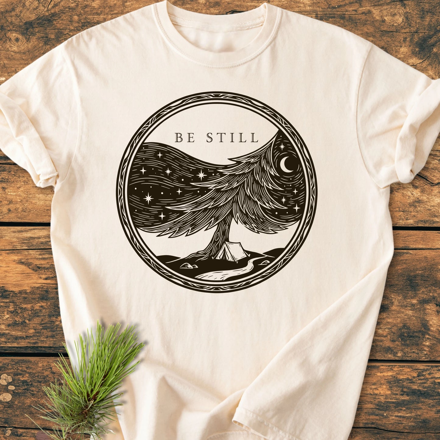 1411 Be Still T-shirt features a woodcut-style pine tree scene with 'BE STILL' text on a cream base, displayed on a rustic wooden surface with a pine sprig.