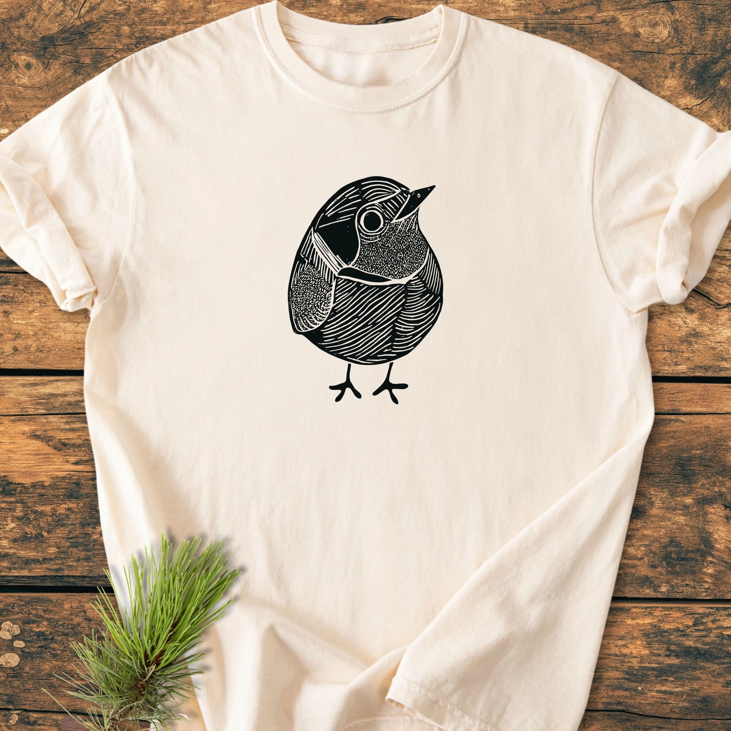 Light-cream T-shirt titled 1433 Little Robin, features a black line-art bird illustration. Laid on a wooden surface with a pine sprig, highlighting its vintage wash and relaxed unisex fit.