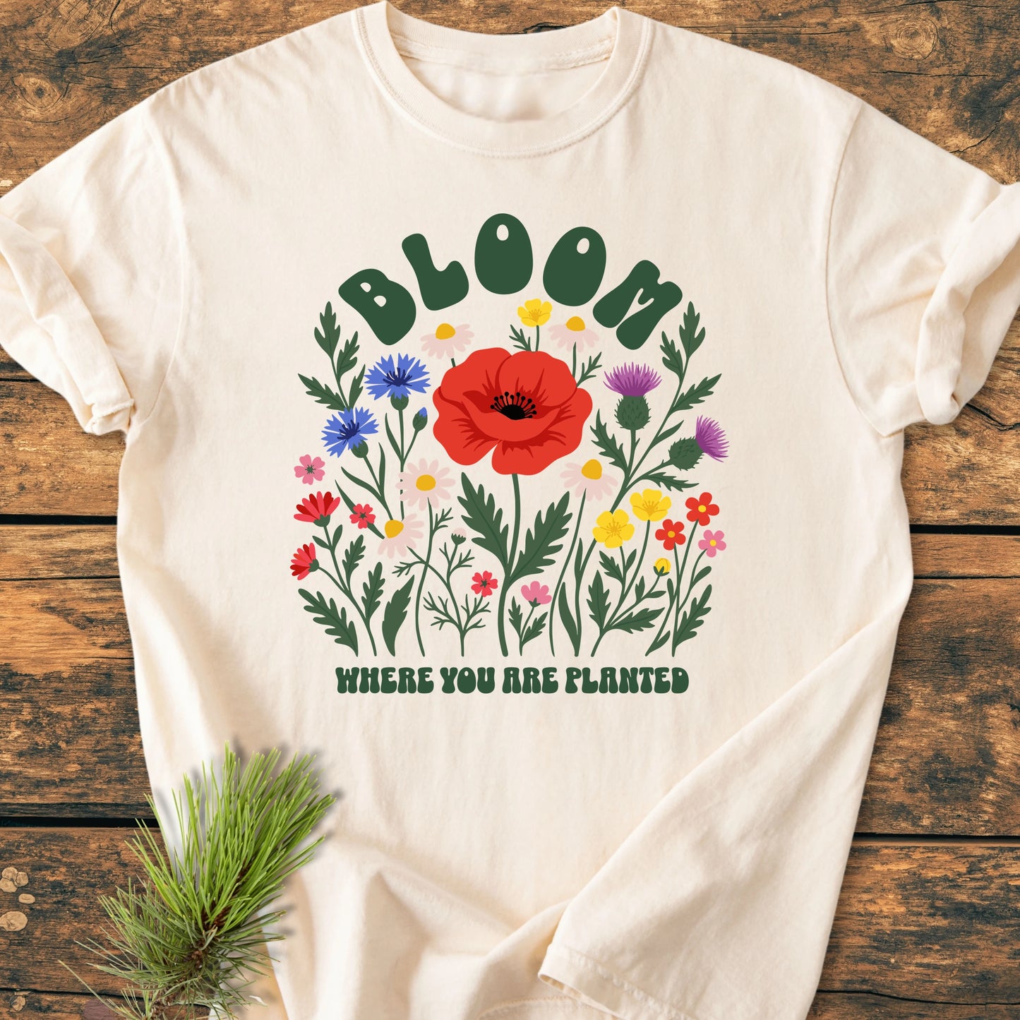 Cream-colored T-shirt, 1403 Bloom Where You Are Planted Base, features a stylized wildflower bouquet with BLOOM and WHERE YOU ARE PLANTED text, laid on a wooden surface with a pine sprig.