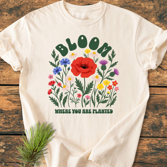 Cream-colored T-shirt, 1403 Bloom Where You Are Planted Base, features a stylized wildflower bouquet with BLOOM and WHERE YOU ARE PLANTED text, laid on a wooden surface with a pine sprig.