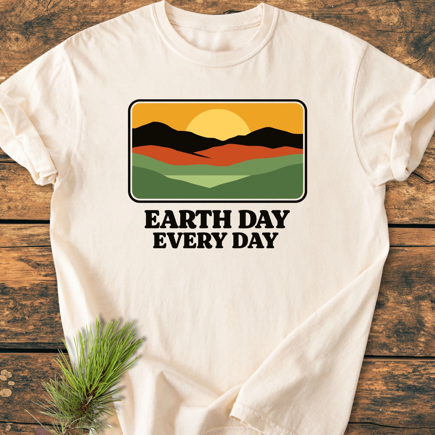 Cream T-shirt on wood, featuring a retro landscape graphic with hills, mountains, and sun. Bold text reads: “EARTH DAY EVERY DAY.” Designed for a relaxed, vintage-wash look and feel.