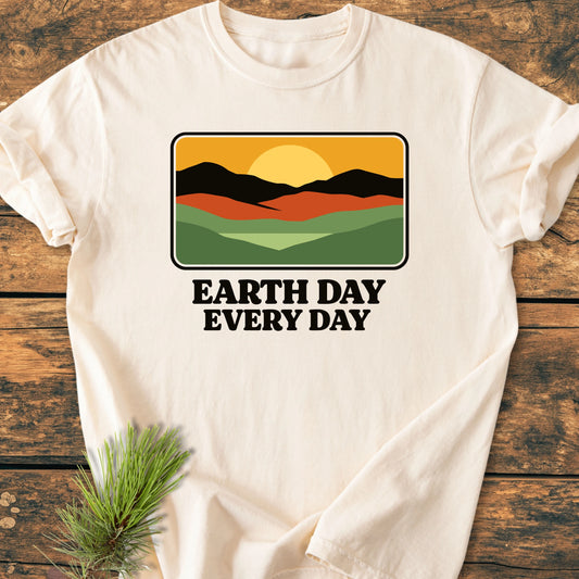 Cream T-shirt on wood, featuring a retro landscape graphic with hills, mountains, and sun. Bold text reads: “EARTH DAY EVERY DAY.” Designed for a relaxed, vintage-wash look and feel.