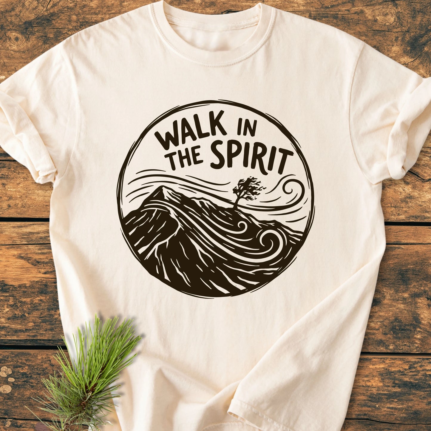 1410 Walk In The Spirit T-shirt on rustic wood, featuring a circular hand-drawn graphic with text, rolling hills, and a windswept tree, embodying the comfort and relaxed fit of garment-dyed cotton.
