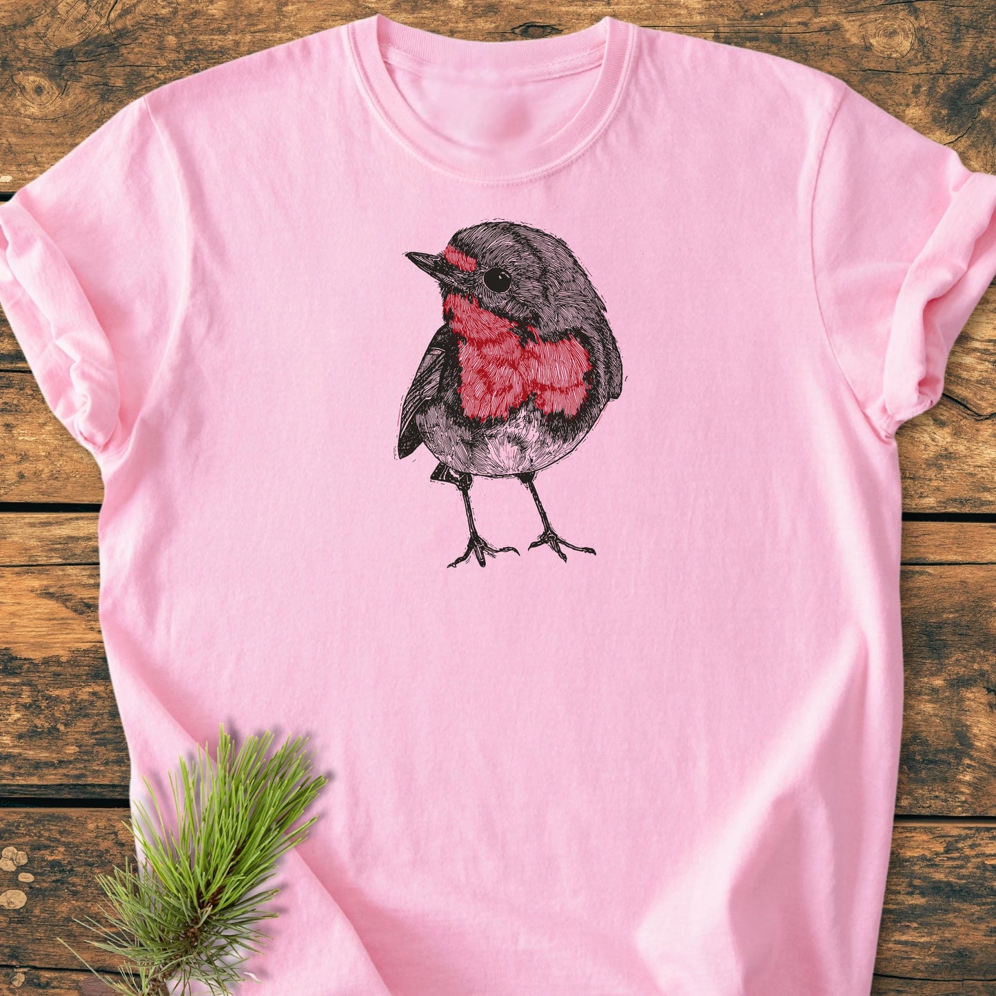 Cute Robin T-shirt features a hand-drawn robin illustration on a soft-pink fabric, laid on a wooden surface, accompanied by a pine branch. Designed for a relaxed, vintage-inspired fit.