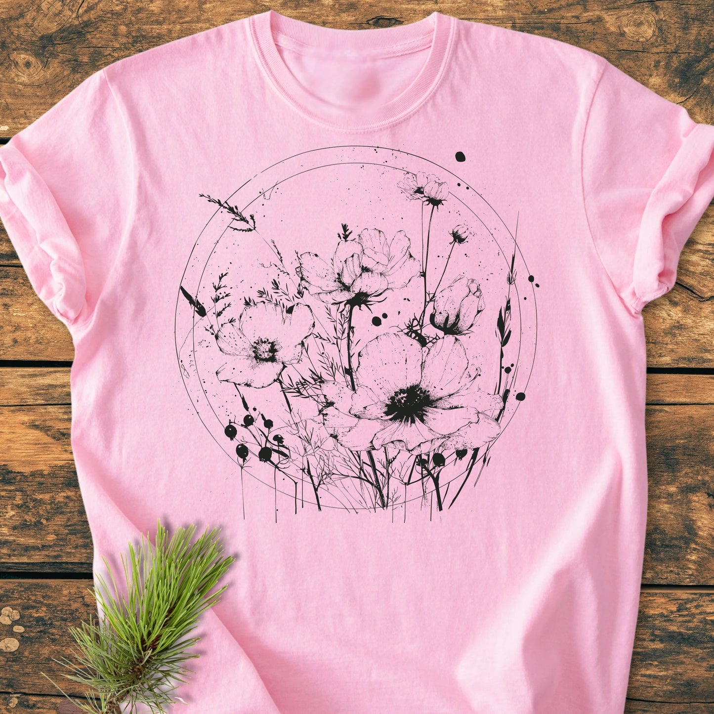 Light-pink T-shirt 391 Wildflower Circle featuring a hand-drawn black wildflower graphic, laid flat on wood. Includes a pine sprig for a natural touch, highlighting the shirt's relaxed, durable design.