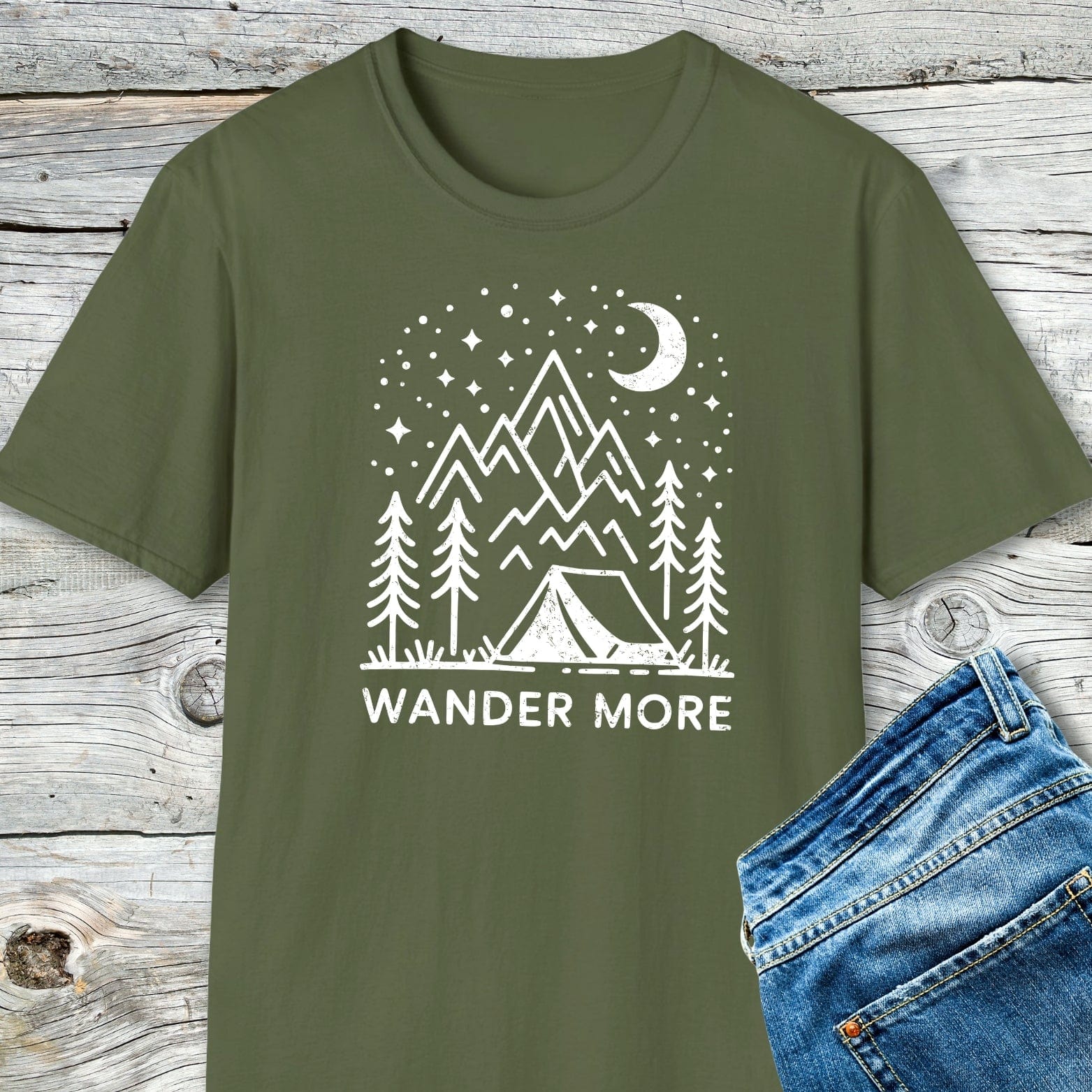 Wander More T-Shirt featuring a camping scene print, laid on wood with jeans; crafted from soft cotton, pre-shrunk, durable, and designed for outdoor enthusiasts.