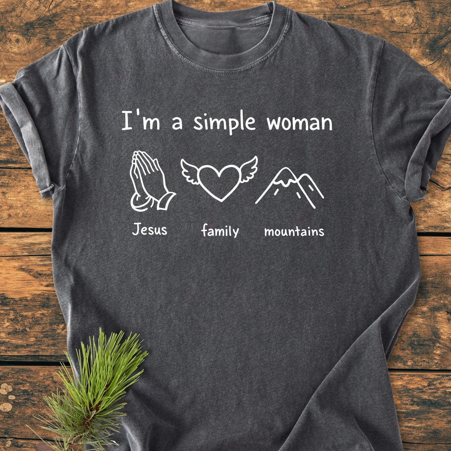 Charcoal-gray T-shirt with “I’m a Simple Woman” printed in white, featuring icons: praying hands labeled “Jesus,” heart with wings labeled “family,” and stylized mountain peak labeled “mountains.”