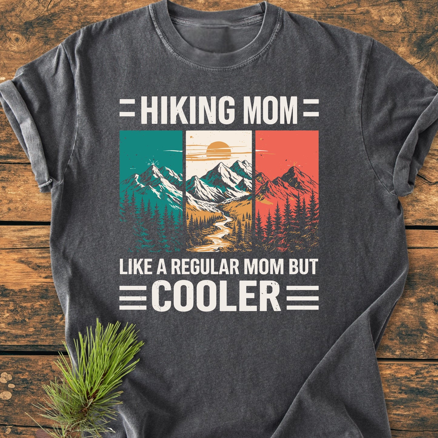 Hiking Mom - Vintage Wash Tee: Dark charcoal-gray T-shirt featuring a mountain-and-forest graphic and playful text, laid flat on wood with a pine sprig accentuating its outdoor theme.