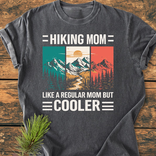 Hiking Mom - Vintage Wash Tee: Dark charcoal-gray T-shirt featuring a mountain-and-forest graphic and playful text, laid flat on wood with a pine sprig accentuating its outdoor theme.
