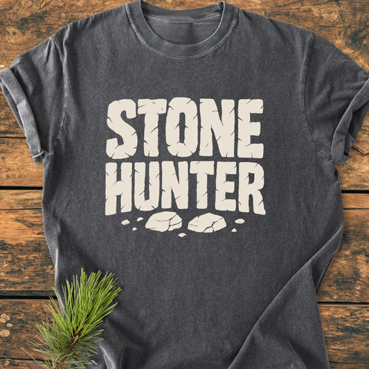 Charcoal-gray 1425 Stone Hunter T-shirt with cracked-stone cream font and stone-shard graphics, laid on rustic wood with pine sprig, exemplifying a vintage-wash, relaxed unisex fit for everyday wear.