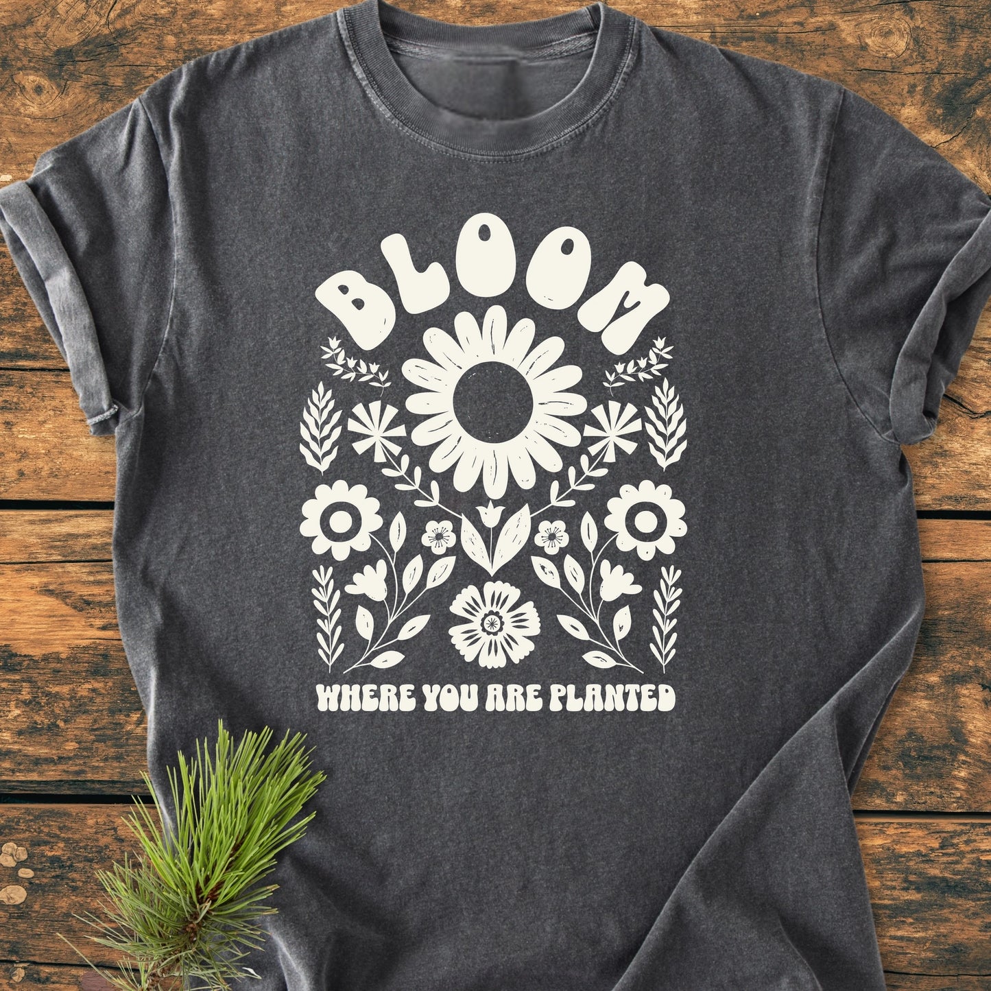 Dark-gray short-sleeve T-shirt featuring a white floral design with BLOOM WHERE YOU ARE PLANTED. Laid flat, highlighting its relaxed fit and vintage wash, ideal for everyday wear.