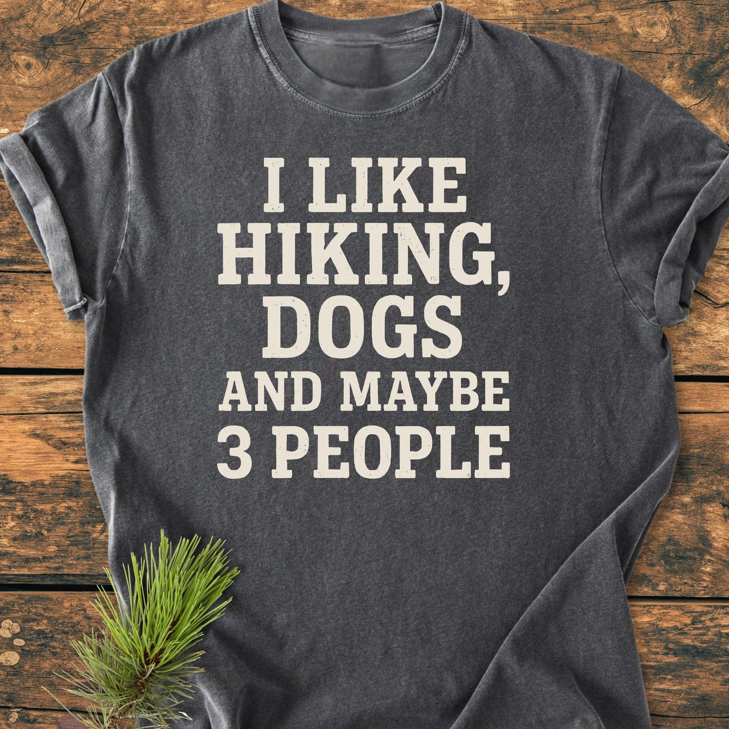 Charcoal-gray T-shirt with I LIKE HIKING, DOGS AND MAYBE 3 PEOPLE text, laid on wood. Rolled sleeves, pine sprig accent. Vintage wash, soft, relaxed unisex fit. Product title: 1431 I Like Hiking And Dogs.