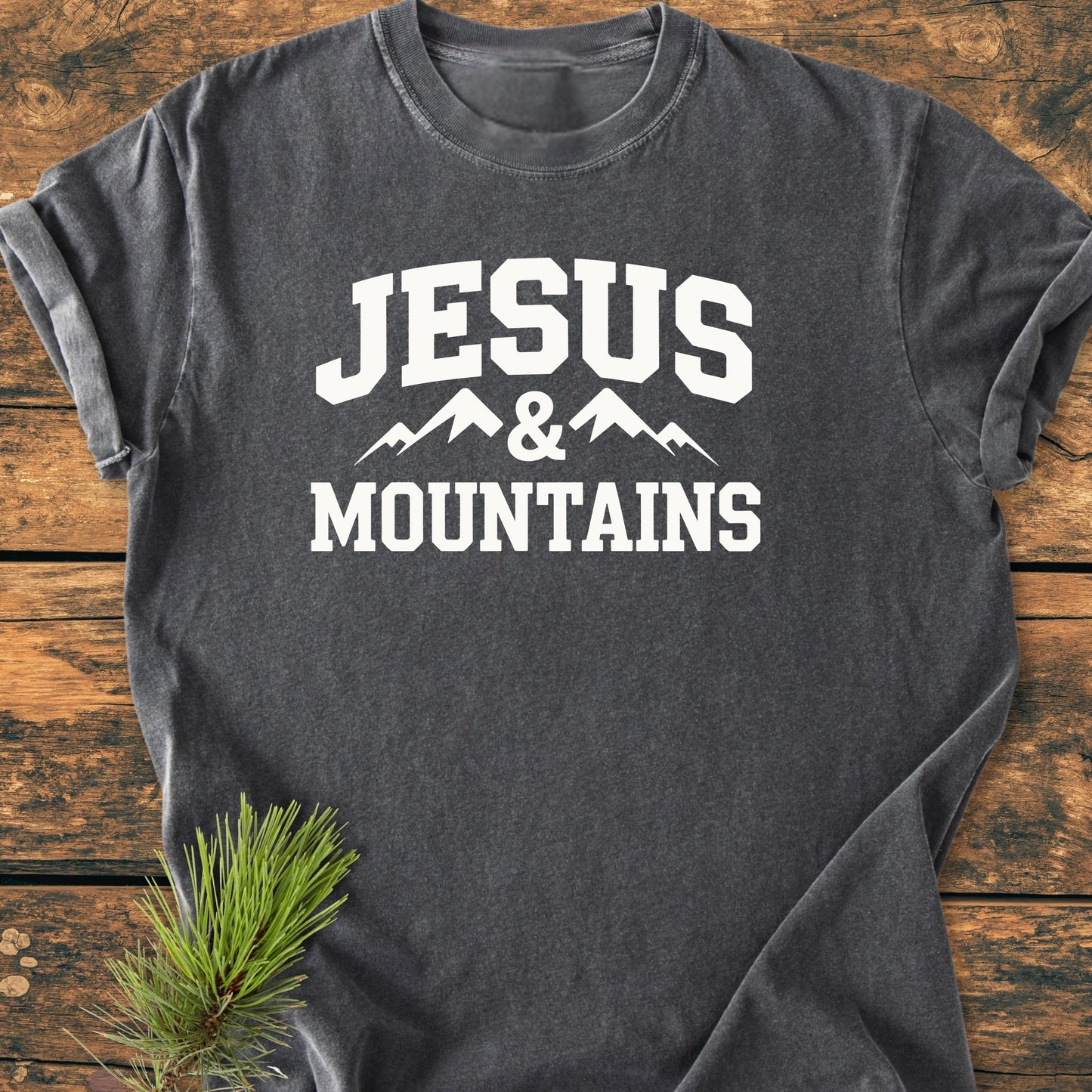Charcoal-gray T-shirt with bold “JESUS & MOUNTAINS” text and mountain graphic, laid flat on wood. Pine branch adds rustic touch, showcasing the vintage wash and relaxed fit.