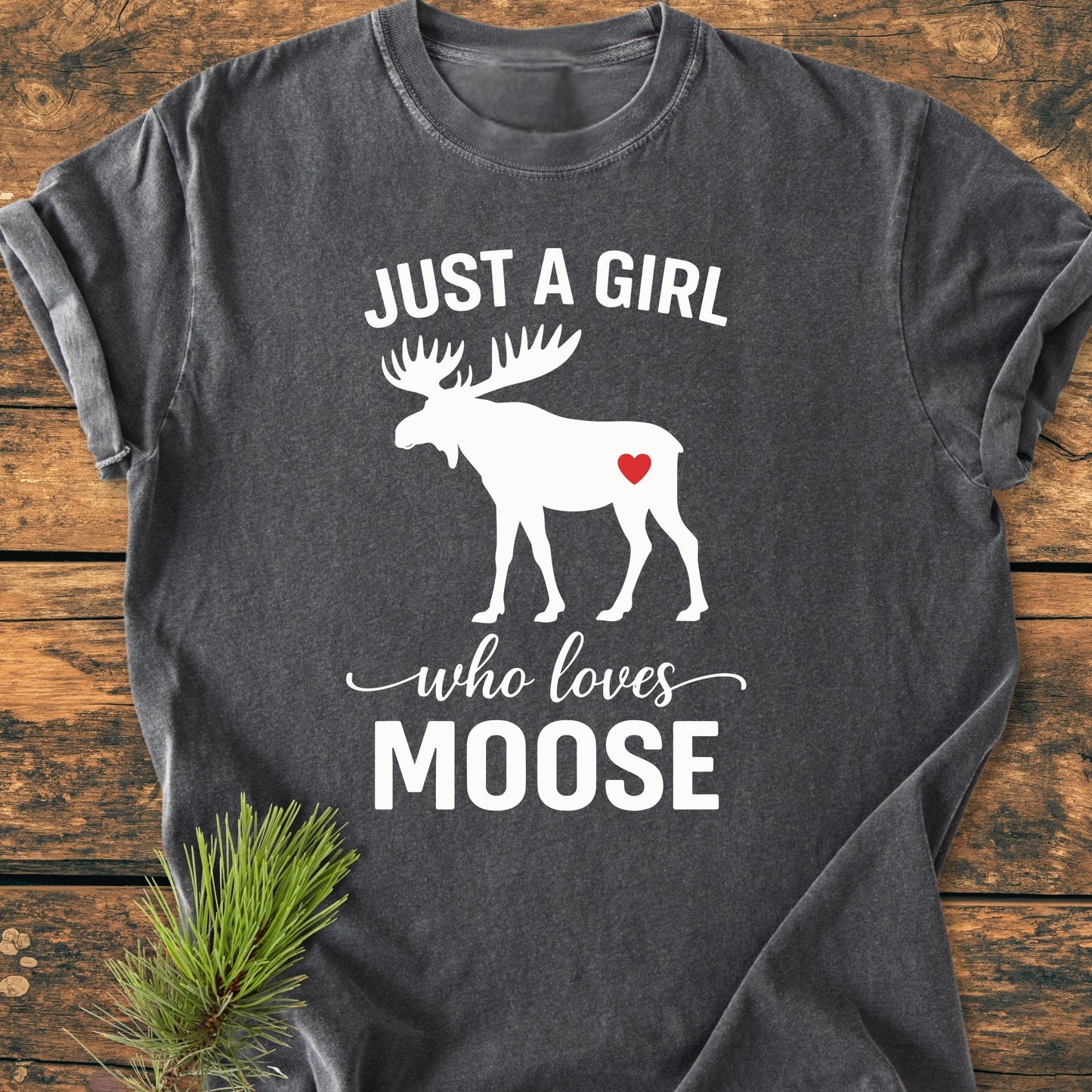 Dark-gray T-shirt featuring 1437 Just A Girl Who Loves Moose slogan with white moose silhouette and red heart, laid flat on wood surface with pine sprig, highlighting its vintage wash softness and relaxed fit.