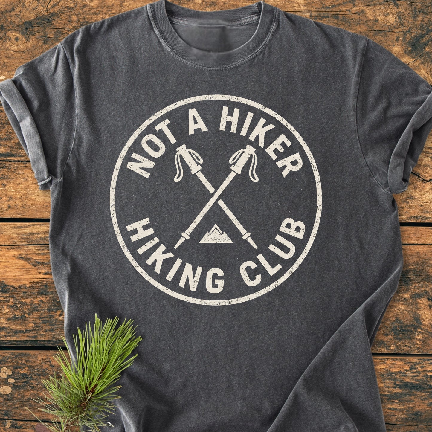 Dark gray 1426 Not A Hiker tee with circular logo featuring crossed hiking poles and mountain silhouette, laid on wood. Includes a nearby pine branch, highlighting its outdoor-inspired design.