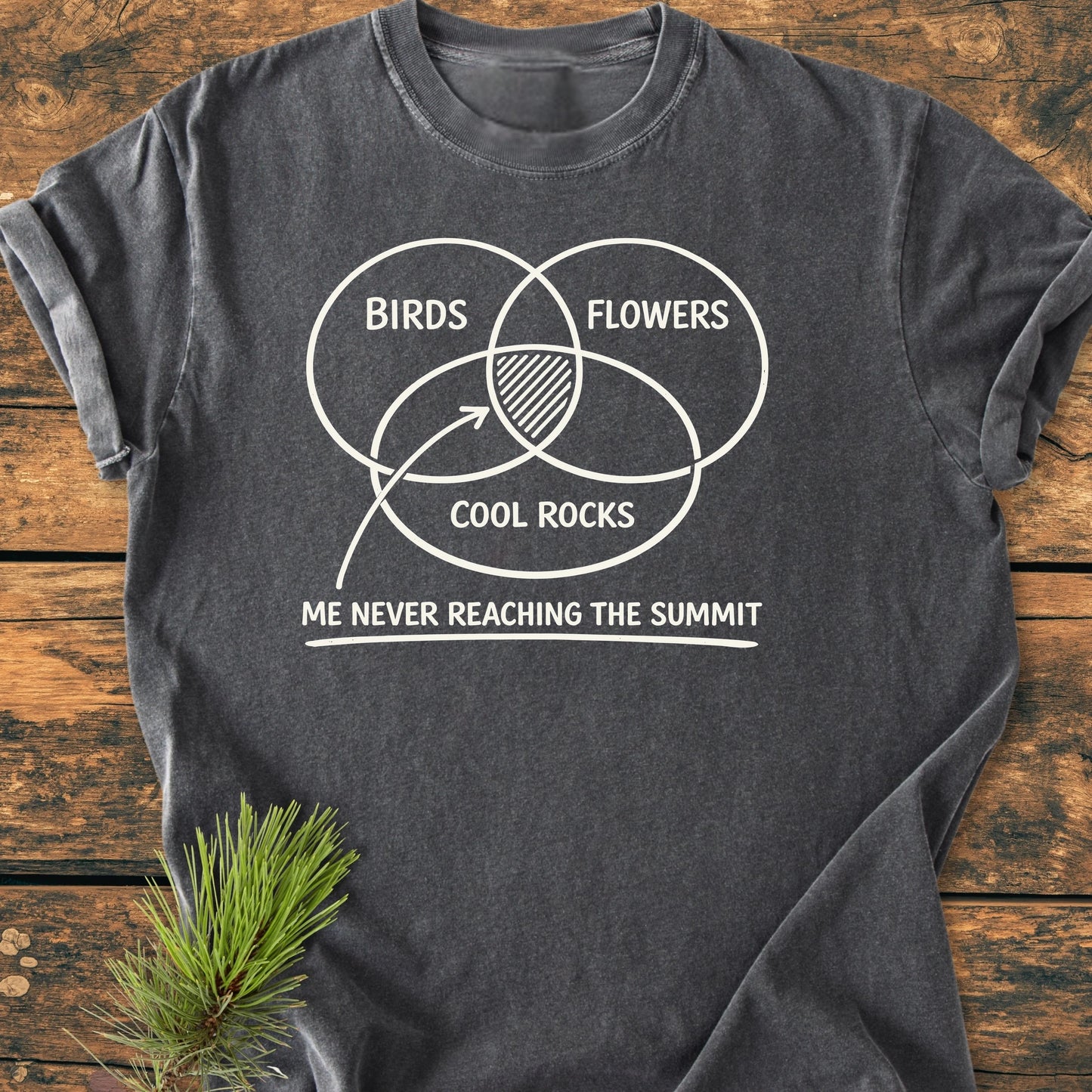 Charcoal-gray 1416 Easily Distracted Hiker T-shirt features a Venn diagram with BIRDS, FLOWERS, and COOL ROCKS, highlighting the center caption ME NEVER REACHING THE SUMMIT, laid beside a pine sprig.