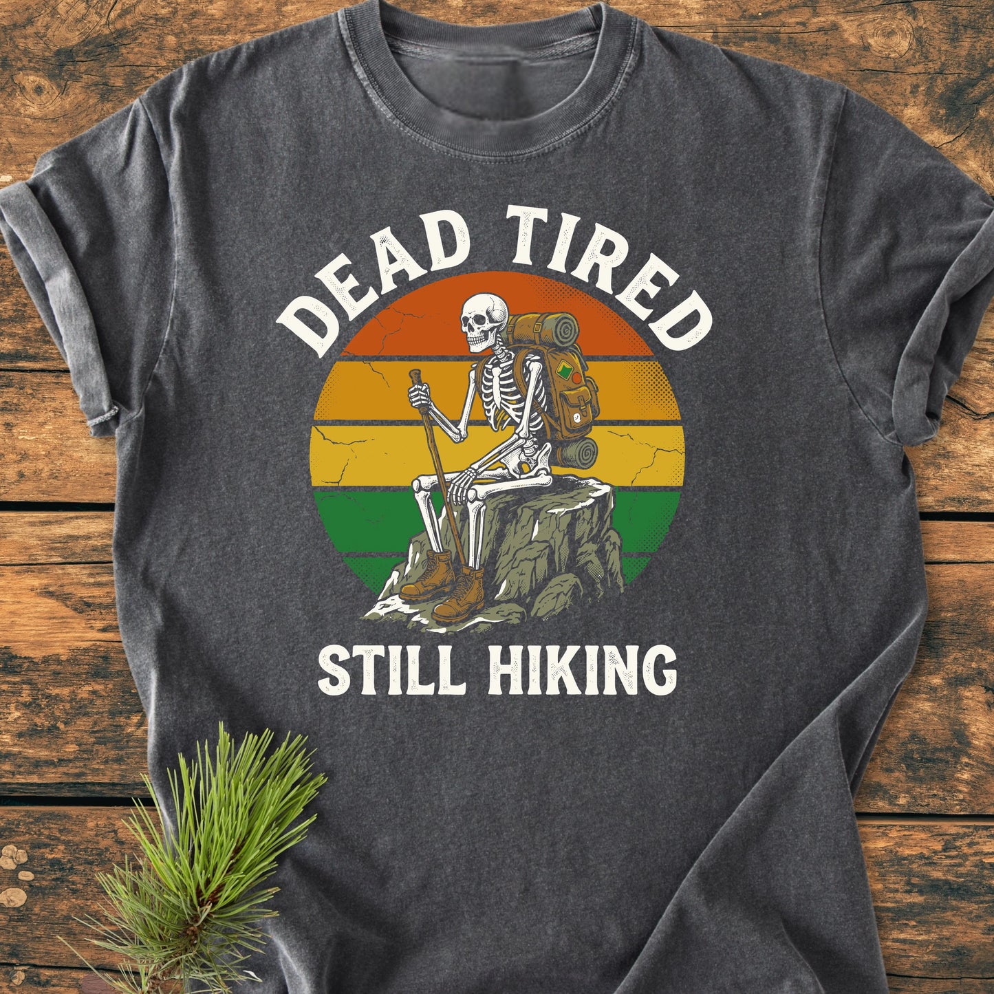 Dead Tired - Vintage Wash Tee features a skeleton graphic with hiking gear, set against a sunset. Laid on wood, the charcoal-gray shirt highlights cozy fabric and relaxed fit, perfect for daily wear.