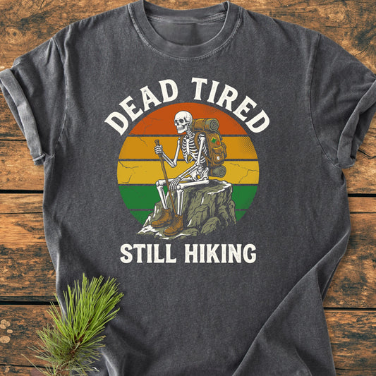 Dead Tired - Vintage Wash Tee features a skeleton graphic with hiking gear, set against a sunset. Laid on wood, the charcoal-gray shirt highlights cozy fabric and relaxed fit, perfect for daily wear.