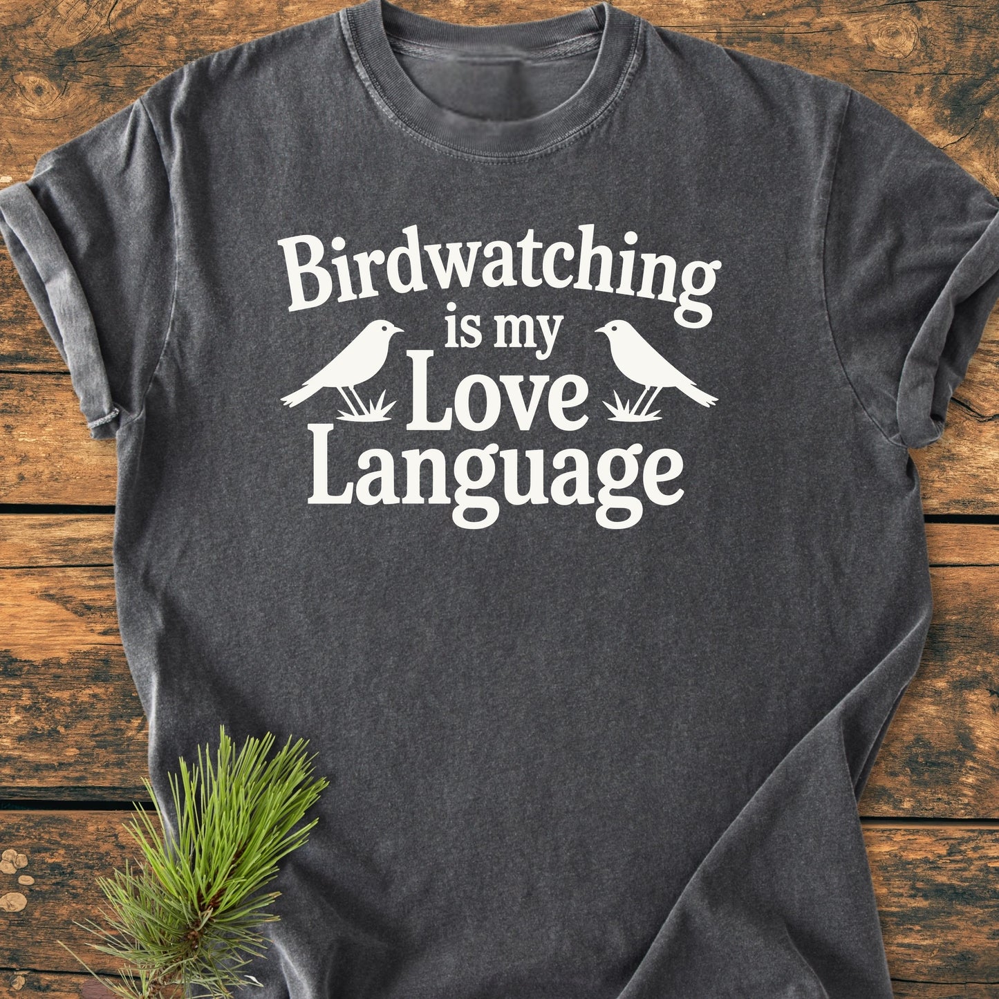 Charcoal-gray T-shirt featuring “Birdwatching is my Love Language” in white, with bird silhouettes and stylized grass, laid flat on wood with a pine sprig nearby.