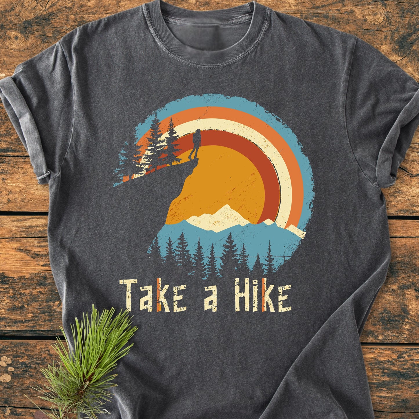 Take A Hike - Vintage Wash Tee featuring a retro outdoors graphic with a hiker and mountains, displayed on wood with a pine sprig, highlighting its relaxed, soft-washed design for everyday wear.