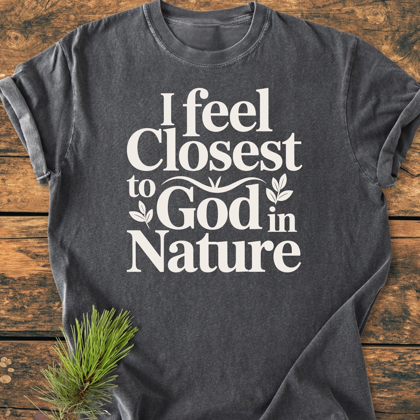 Charcoal-gray 1427 Closest To God - Vintage Wash Tee with I feel Closest to God in Nature slogan and leaf motifs, displayed on wood, paired with a pine branch accentuating its nature-inspired theme.