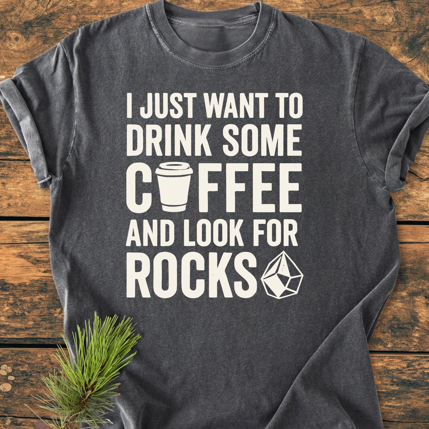 Heather-gray T-shirt titled 1404 Drink Coffee And Look For Rocks Base with a playful coffee and rock theme, laid flat on wood with a pine sprig accentuating its cozy, relaxed fit design.