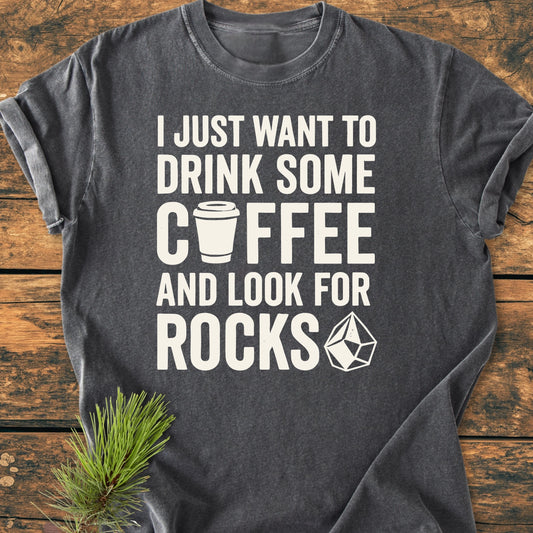 Heather-gray T-shirt titled 1404 Drink Coffee And Look For Rocks Base with a playful coffee and rock theme, laid flat on wood with a pine sprig accentuating its cozy, relaxed fit design.